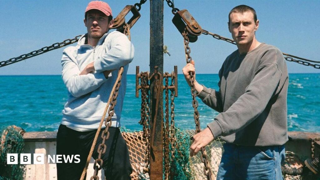 A scene from the film in which George MacKay and Callum Turner are standing on a fishing boat while at sea on a sunny day. MacKay is holding on to a chain fed into a pulley while Turner leans on a chain with his arms crossed. They are both looking steadily ahead.