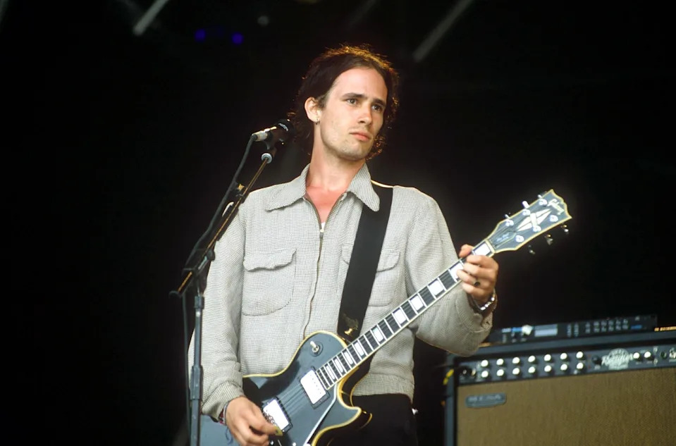 Jeff Buckley playing an electric guitar on stage, wearing a casual zip-up jacket, singing into a microphone
