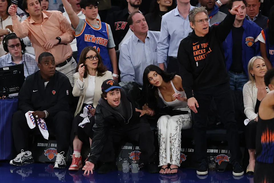 People sitting courtside at a basketball game, reacting to the action on the court. Some are standing, pointing, or cheering