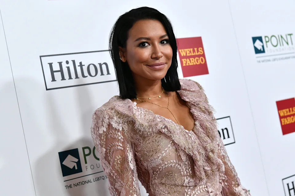 Naya Rivera in a lace dress poses on a red carpet at an event with sponsor logos in the background