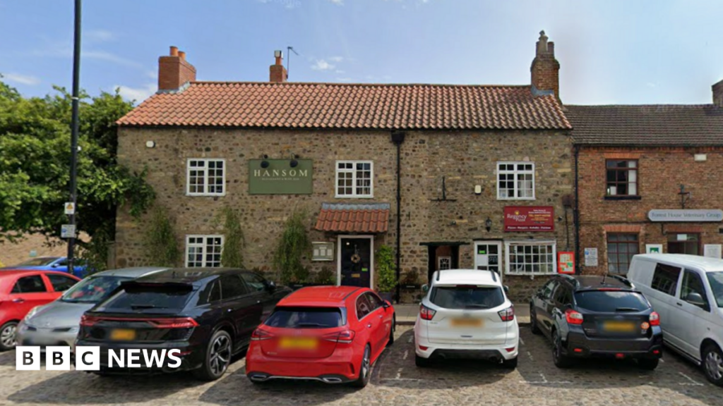 A Google Maps image of the Hansom Restaurant in Bedale, a large stone building that neighbours a pizza takeaway. The building has five large windows with white frames, and a black drain pipe down the middle dividing the two properties and their front doors. Hansom has a tiled awning above their black door, and there are seven cars parked along the street in front of it. The property has a chimney, a tree growing directly next to the restaurant, and greenery is growing up the bricks. There is also a street light off to the left.