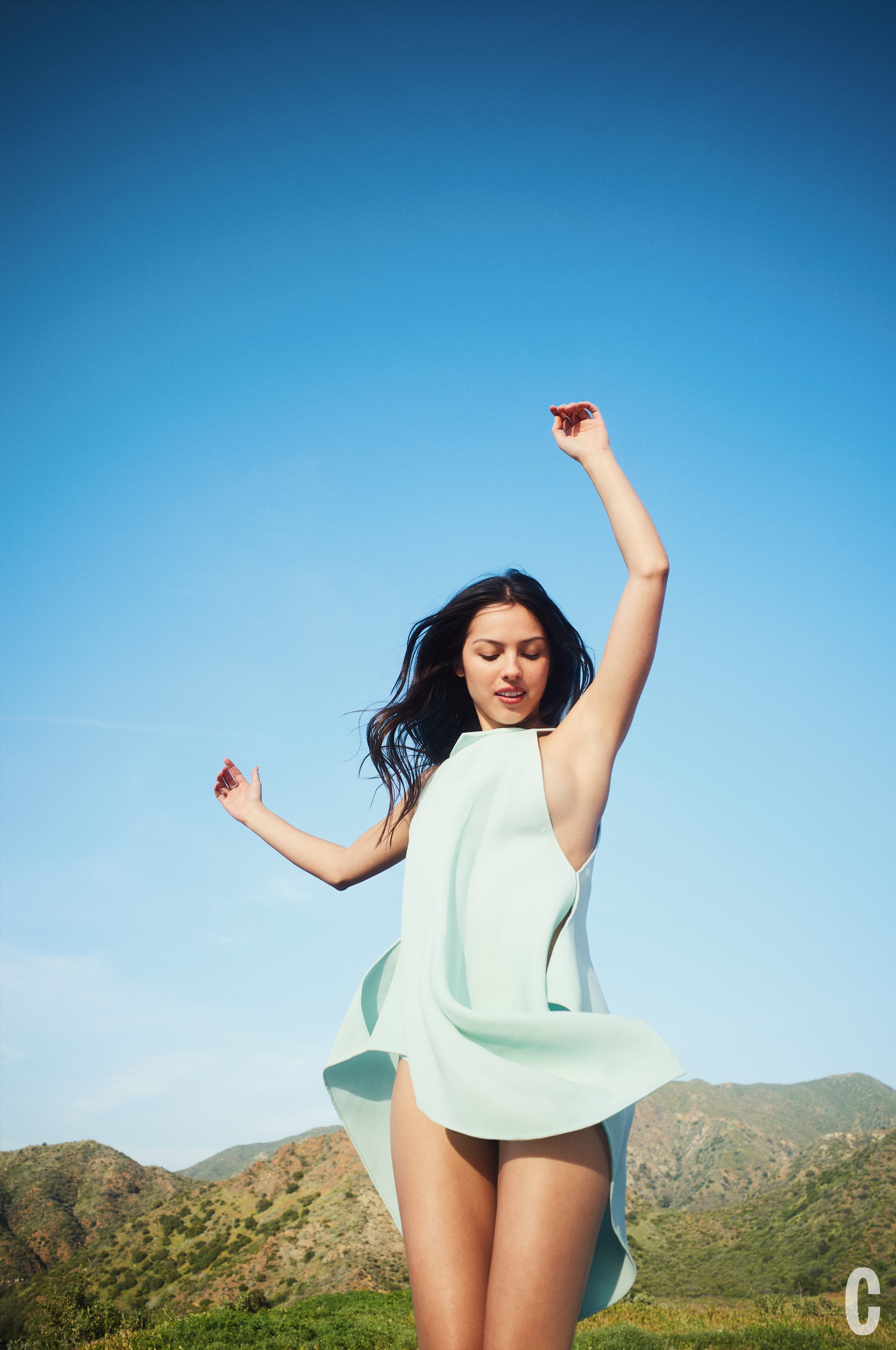 A person joyfully dancing outdoors in a flowing mint green outfit against a clear blue sky.
