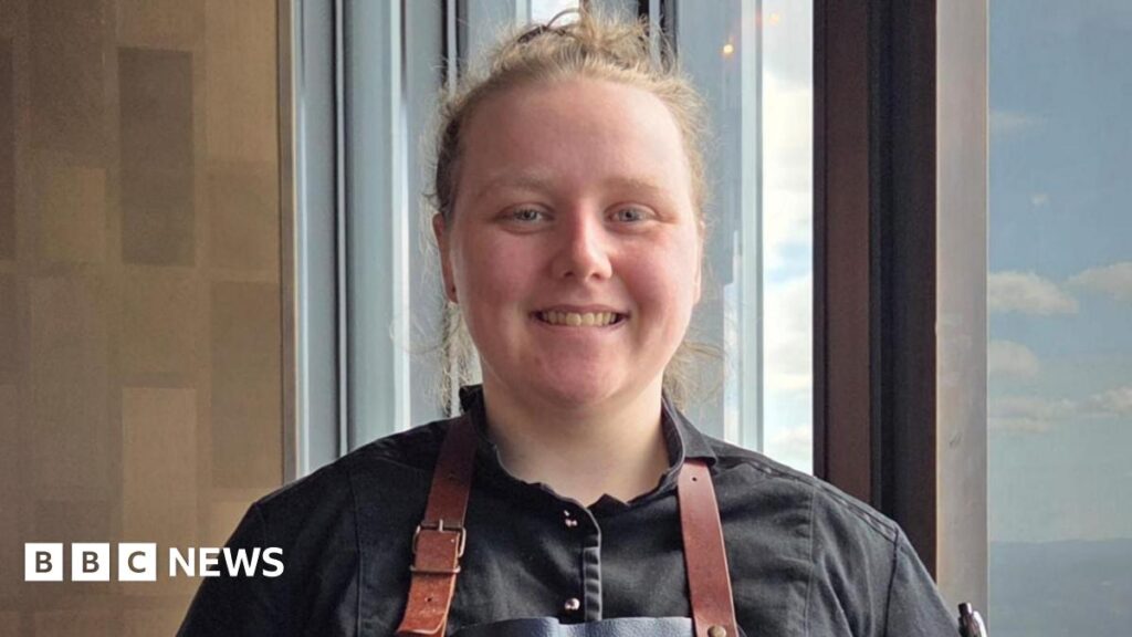 Holly smiles at the camera and has her blonde hair tied in a bun. She wears a black shirt and a brown apron.