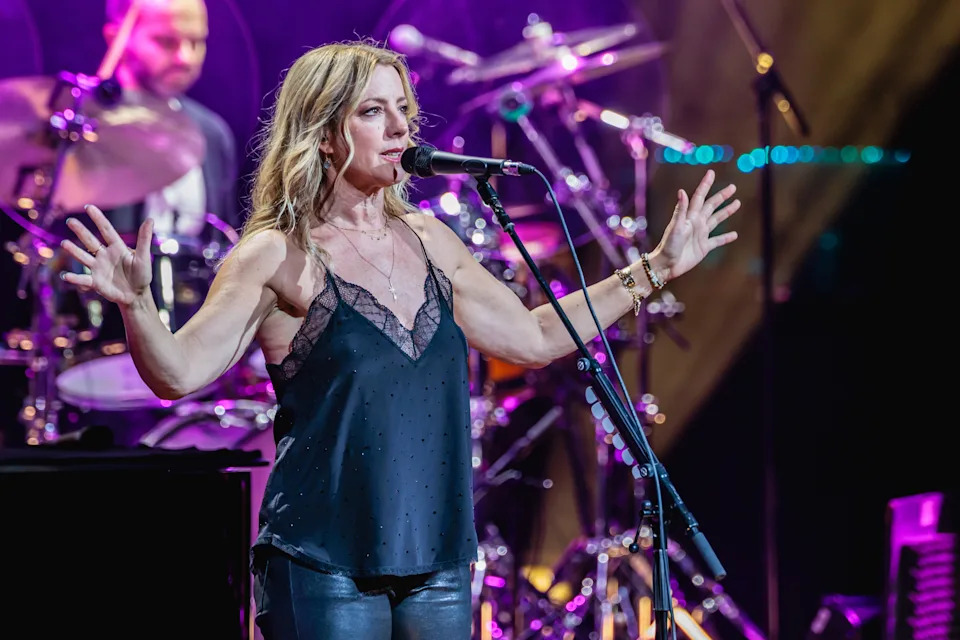 Woman performing on stage in a sleeveless top and leather pants, with a band playing in the background