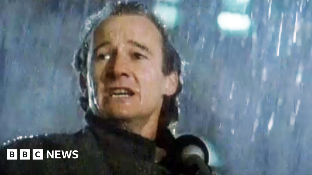 David Hayman in 1986, with long hair and in an overcoat, is standing in heavy rain speaking into a microphone. He looks passionate and serious.