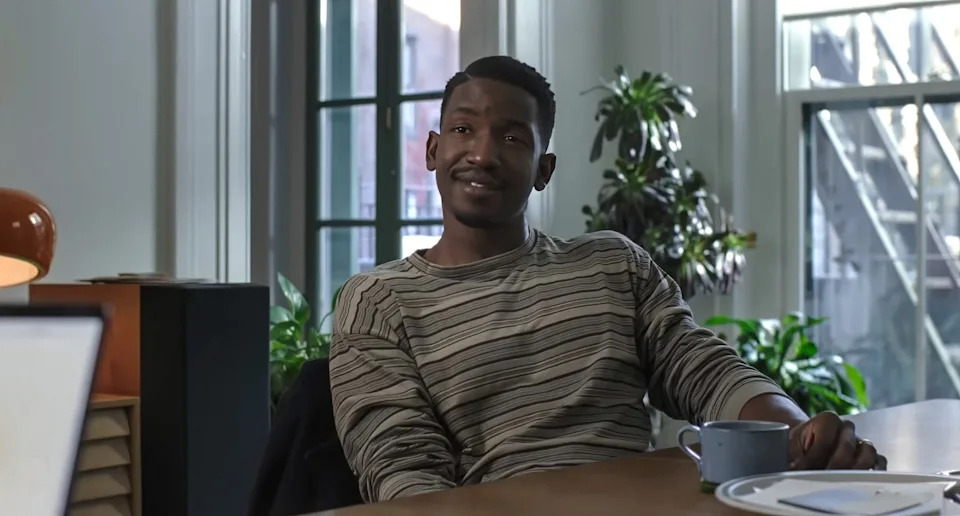 Person sitting at a table in a casual striped shirt, smiling slightly, with plants and a window in the background