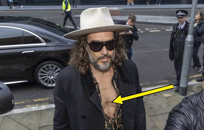 Russell Brand, wearing a wide-brim hat and unbuttoned shirt under a black coat, walks outdoors, surrounded by photographers and onlookers, an arrow points at a cross dangling on his bare chest between buttons opened nearly to his navel