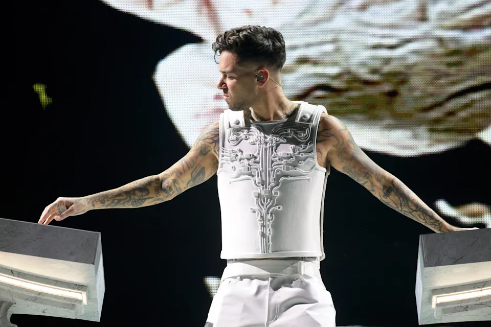 Anyma on stage wearing a detailed armored vest and pants, surrounded by equipment, in an artistic pose during a live performance