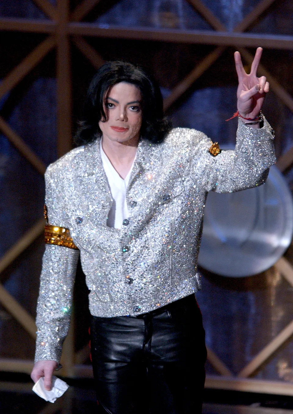 Person in a sparkling jacket and leather pants making a peace sign gesture at a celebrity event