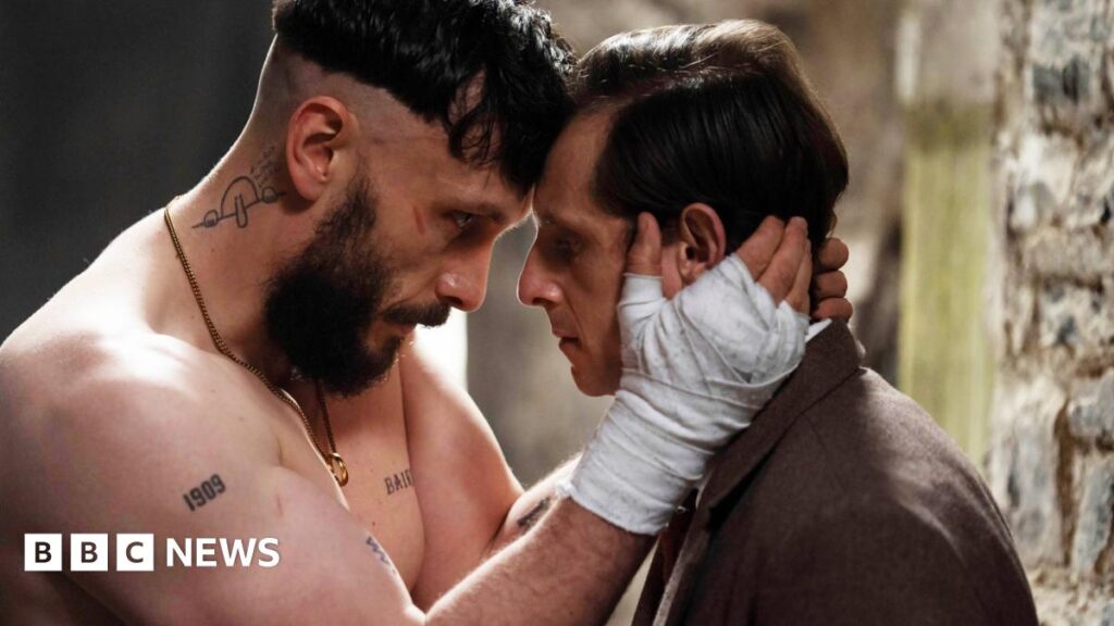 A man with a beard and bandages on his hands holds the face of a man wearing a suit.