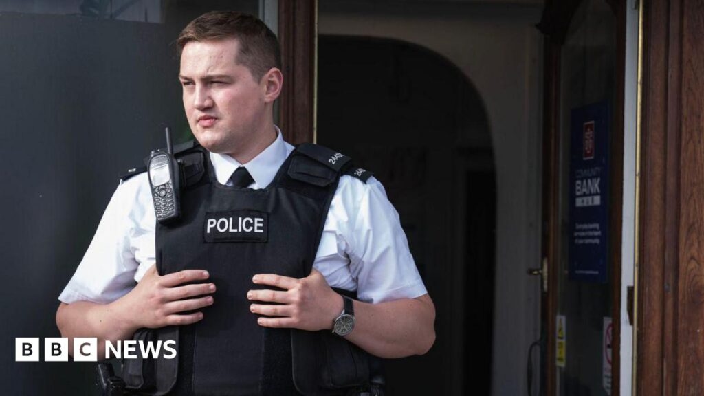 Finnian Garbutt, a man with dark hair, wearing a white shirt with black tie and a black support vest with the words police on it. He is wearing a black watch on his left wrist and there is a brown wooden door in the background.