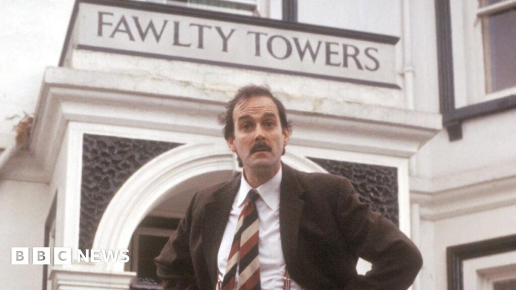 A man with a moustache wearing a suit in front of a hotel saying Fawlty Towers