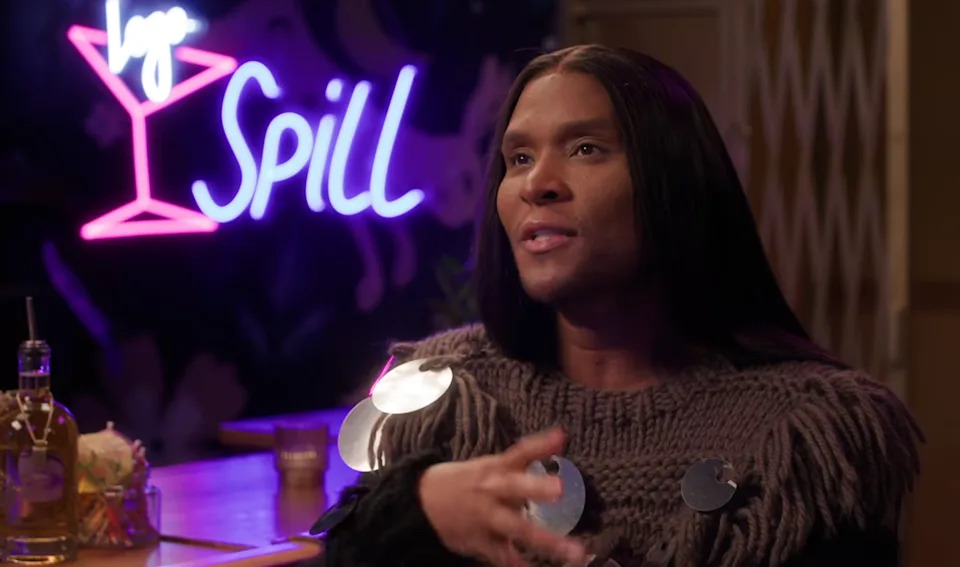 Person speaking in a bar setting, wearing a textured sweater with reflective circular accents. Neon sign in background says "Spill."