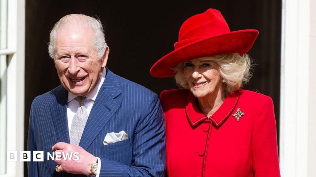 King Charles smiles and looks straight ahead. He is wearing a pinstripe navy suit, a white shirt, a silver tie and a silver pocket square. His left hand is lifted up slightly and he has a black and gold watch on it. His pinky finger on his left hand has a large gold ring. On his left side is Queen Camilla. She is wearing a bright red blazer with a cross-shaped broach. She is wearing a red hat that matches the blazer. She is also looking straight ahead and smiling.
