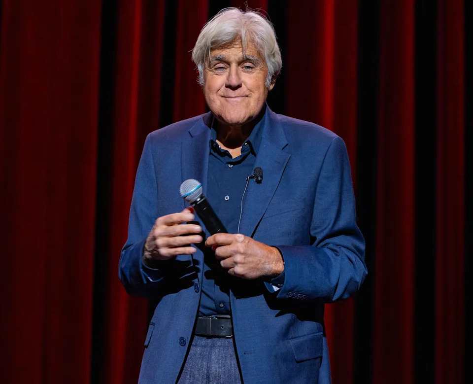 Person in a classic blue suit holding a microphone, standing against a curtain backdrop