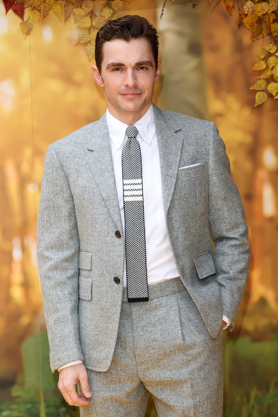 Man in a tailored gray suit, white shirt, and patterned tie, standing with a neutral expression at a formal event