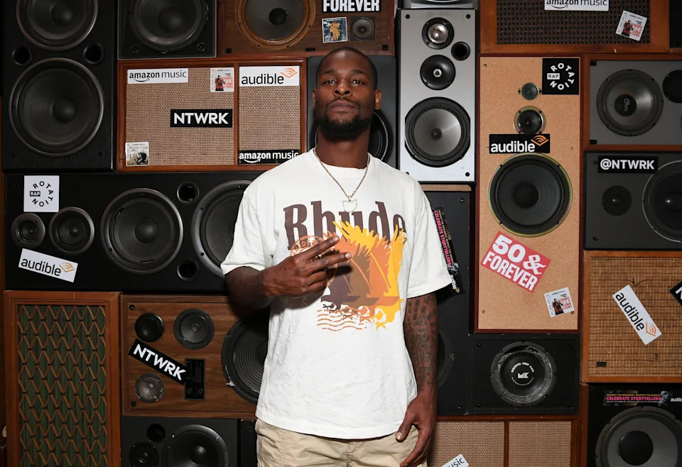 Bell posing in front of a wall of speakers, wearing a graphic t-shirt with "Rhude" text and light shorts, at an audio-themed event
