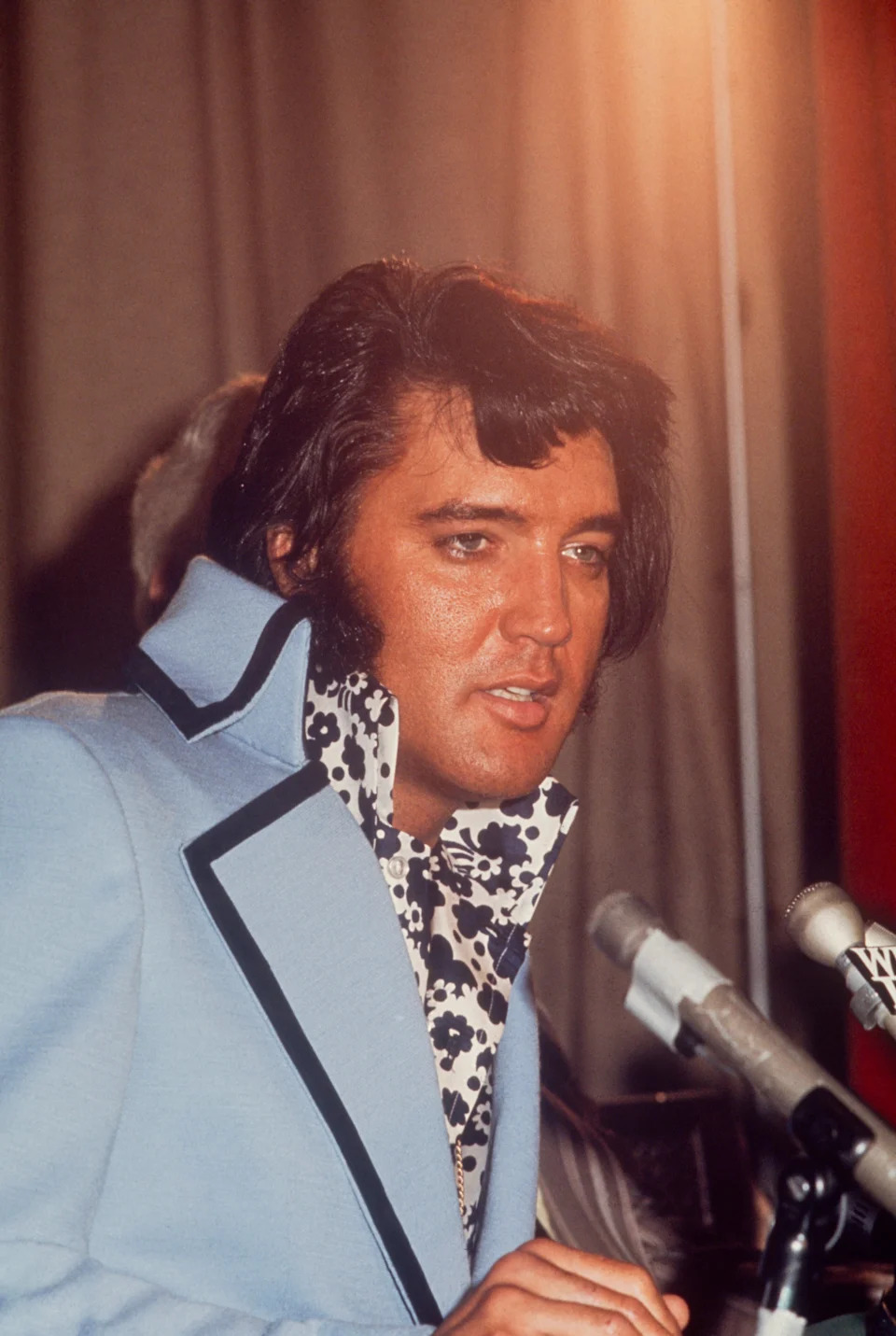 Person with styled hair in a high-collared jacket and patterned shirt speaks at a microphone, appearing at an event or press conference