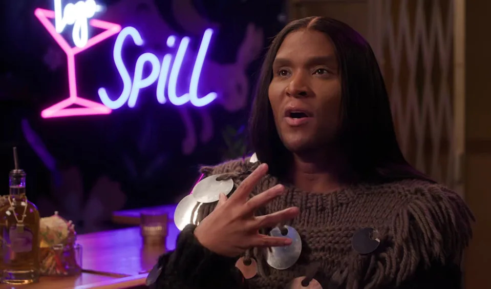 A person sits at a bar with a sign reading "Spill" in the background, wearing a sweater with large decorative discs, gesturing expressively