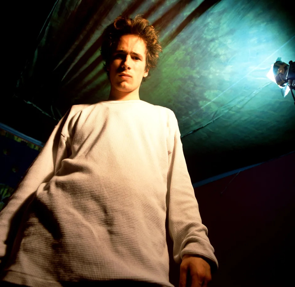 Jeff Buckley standing under a spotlight, wearing a loose-fitting long-sleeve shirt, looking down at the camera