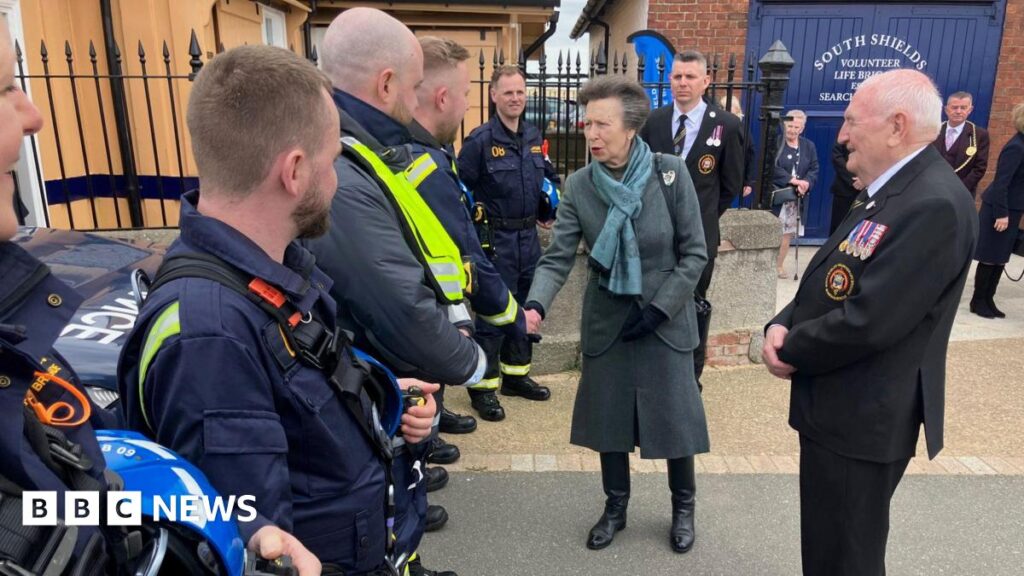 The Princess Royal with her hair tied back. She has a long grey coat, a blue scarf, black gloves, black trousers and black boots. She is shaking hands with members of the South Shields Volunteer Life Brigade in their blue uniforms. There are also dignitaries in uniform. There is a brick building in the background with blue doors.
