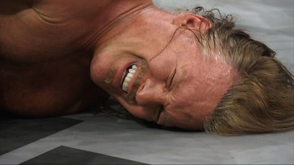 A wrestler lies on the mat with a pained expression, showcasing the aftermath of a match. His long hair and muscular build indicate his experience in the ring.