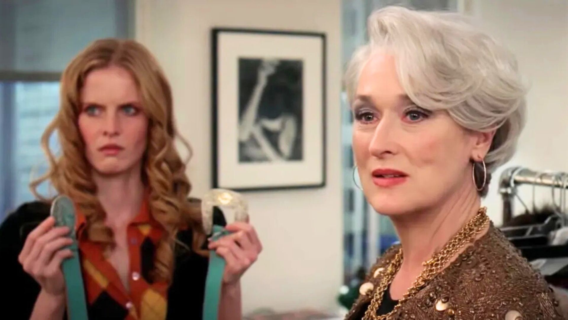 Meryl Streep purses her lips with a woman holding up two cerulean belts behind her