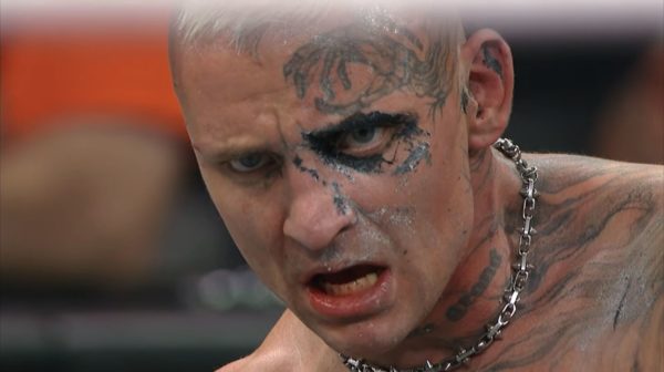 A close-up of a wrestler with a fierce expression, displaying striking facial tattoos and body art. The wrestler has a chain around his neck and appears to be in a high-stakes moment after a match.