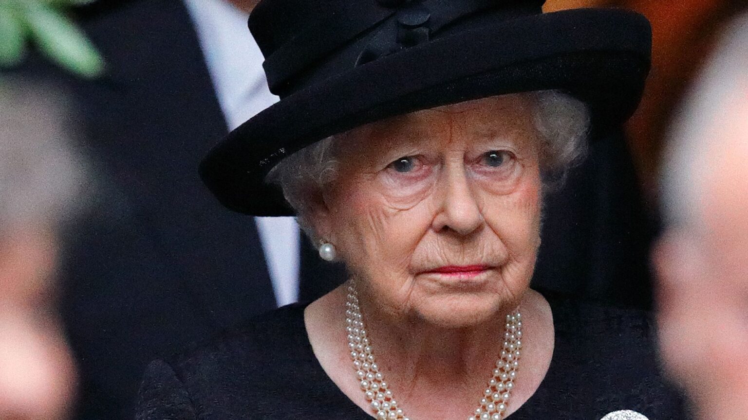 Queen Elizabeth II wears a black dress and matching hat and looks very sad while wearing a string of pearls