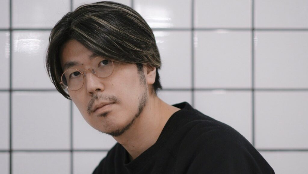 Genki Kawamura interview portrait. Asian man with glasses and highlighted hair in front of tile background.