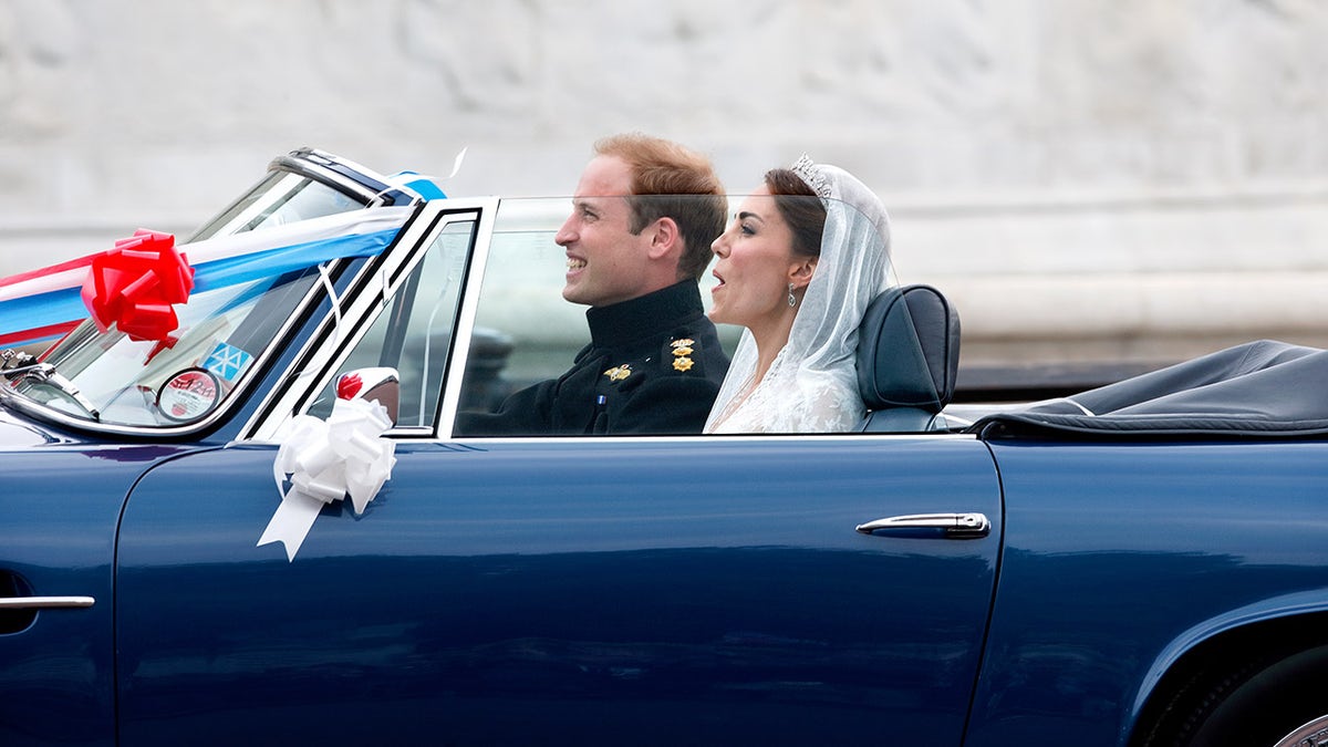 Prince William and Kate Middleton driving a blue Aston Martin convertible decorated with ribbons and balloons.