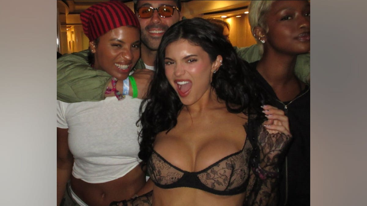 Kylie Jenner posing with a group of friends at an outdoor festival; she is wearing a sheer black long-sleeved top with a built-in lace bra design.