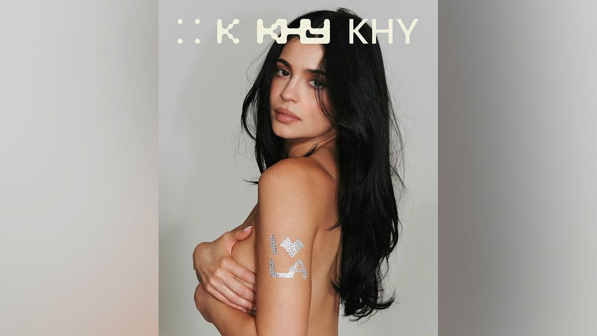 Kylie Jenner posing topless with her arms covering her chest, showcasing a sparkling rhinestone tattoo on her bicep that reads 