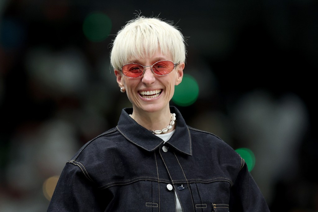 Megan Rapinoe headshot, smiling in pink glasses and a dark denim jacket