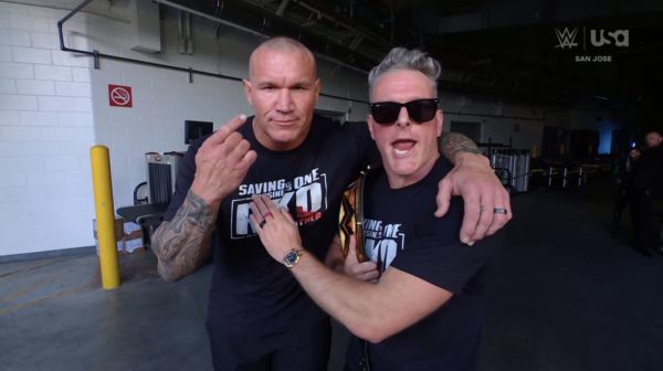 Randy Orton and Pat McAfee pose closely together in a backstage area, with Orton raising one finger, while McAfee has an excited expression and holds a WWE Championship belt. Both wear matching black T-shirts featuring the phrase 'Saving is One' and 'RKO'.