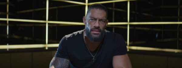 Roman Reigns, a professional wrestler, sits in front of an illuminated backdrop, looking directly at the viewer with an intense expression.