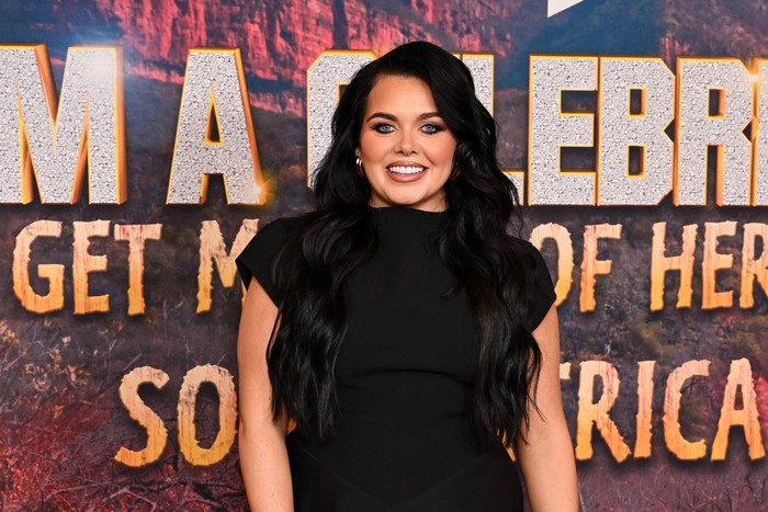 I'm-a-Celeb-South-Africa-2026 Scarlett Mofatt smiling ahead. She wears a black dress and stands in front of a 'I'm a Celebrity Get Me Out of Here South Africa' promotion board