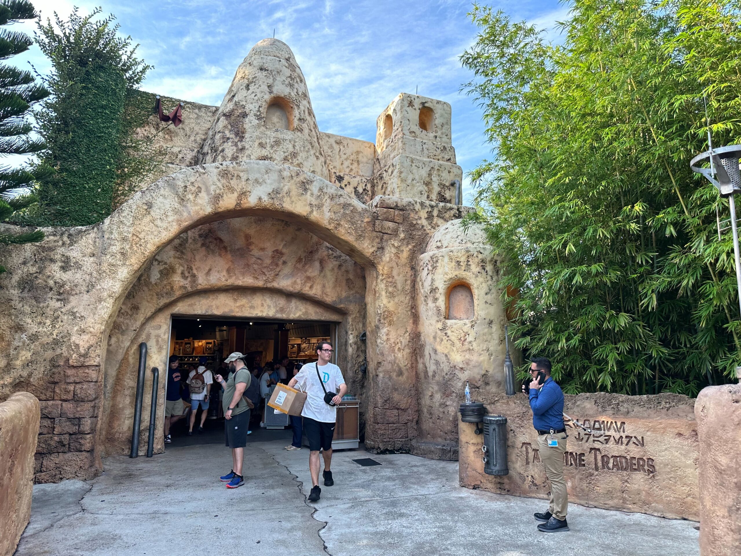 People walking near a stone-built shop entrance at a theme park, with one person carrying a box. The surrounding area has bamboo plants and the sky is clear.