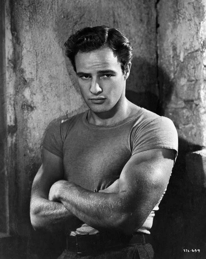 Marlon Brando with arms crossed, wearing a fitted t-shirt. The expression is serious, with a focused gaze. Background is plain and textured