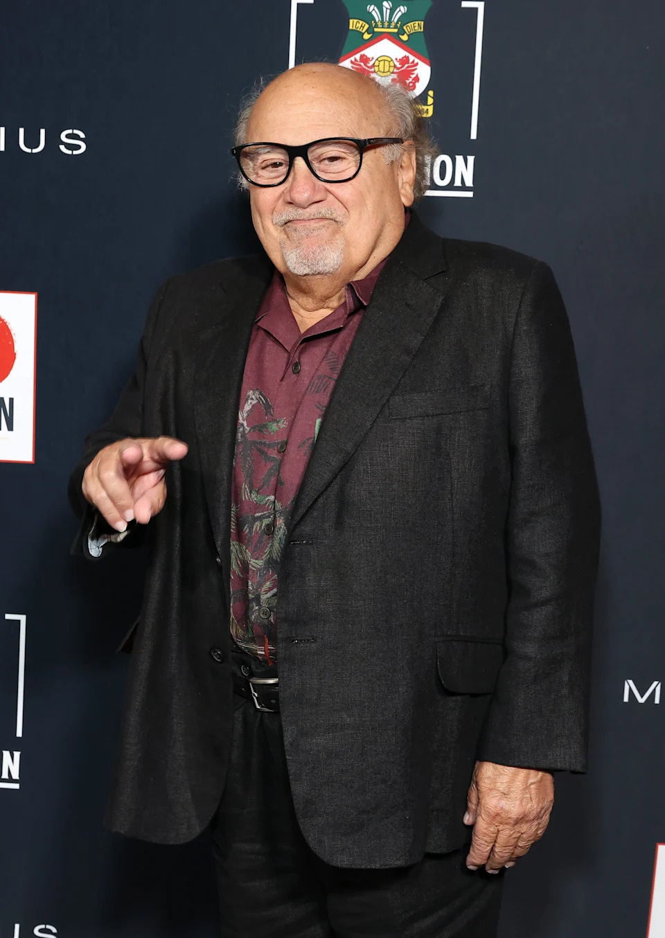 A celebrity wearing a patterned shirt under a tailored suit, posing on the red carpet at a formal event, making a gesture with one hand