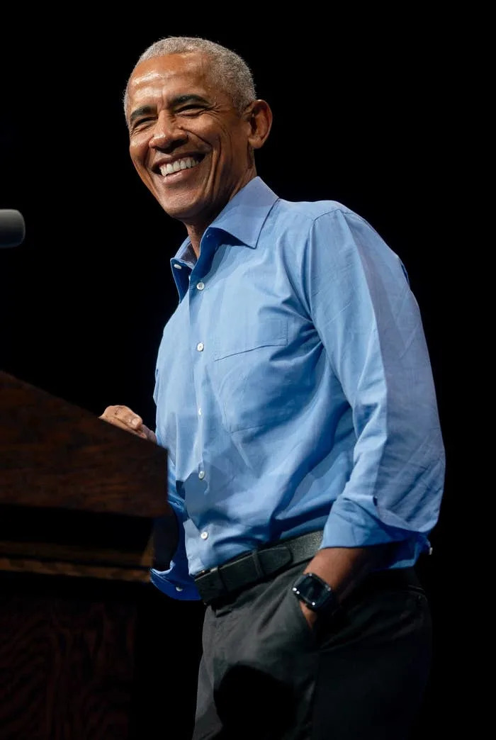 A person in a blue shirt smiles while speaking at a podium with a sign about voting. Another person in a suit is seated nearby, also smiling