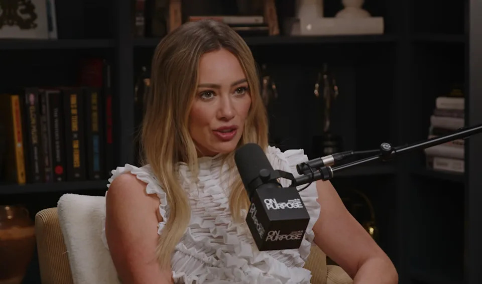 Person in ruffled top speaking into microphone, seated in a studio with bookshelves in the background