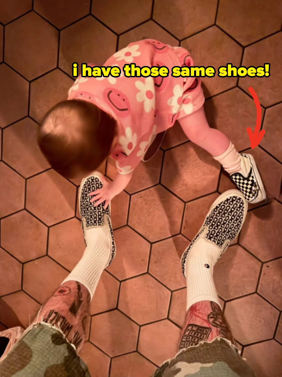 Tattooed adult wearing patterned slip-on shoes and shorts stands with a baby in a floral outfit, bending down on a tiled floor