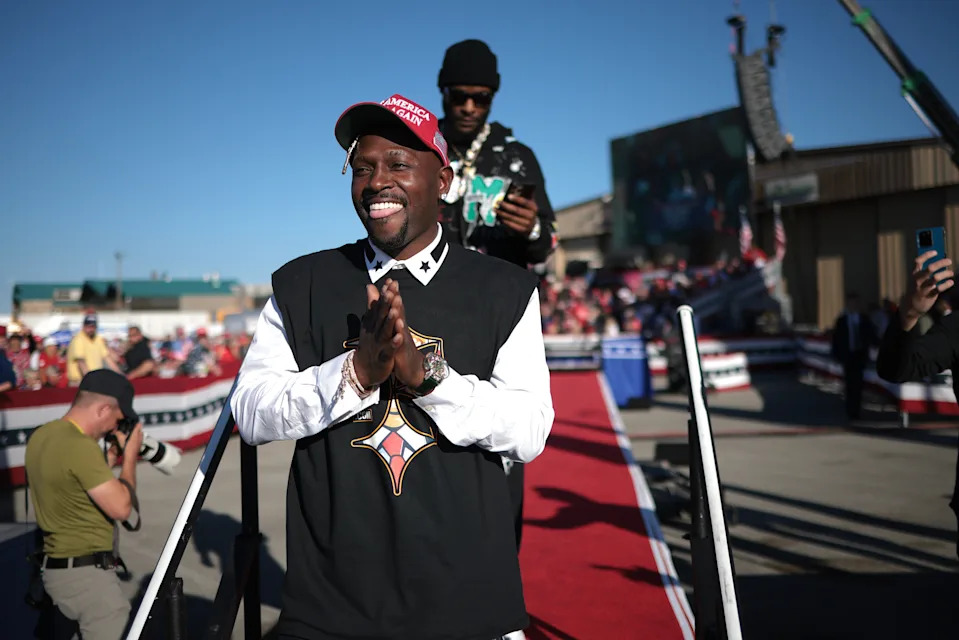 Antonio Brown on stage clapping, wearing a cap and patterned sweater over a white shirt. Another person stands behind him. Audience and flags in background