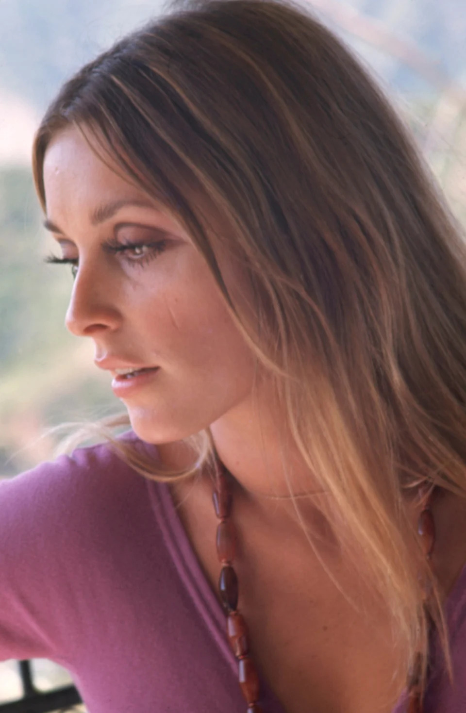 Sharon Tate with long hair and necklace gazes thoughtfully to the side, wearing a casual top