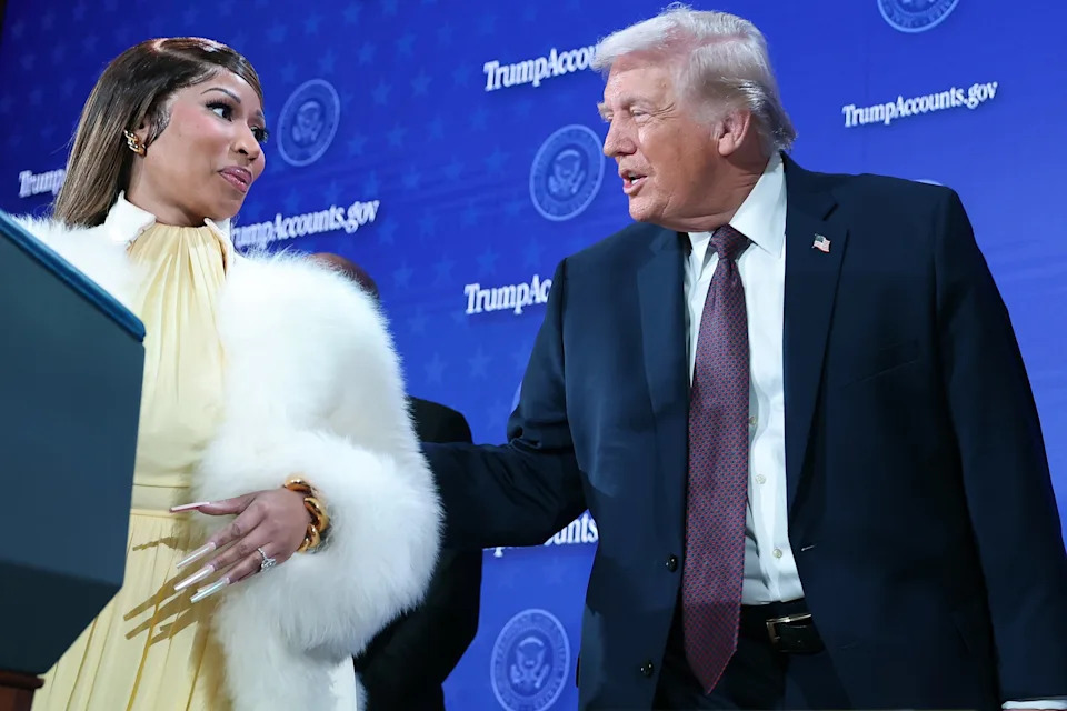 Nicki Minaj in a glamorous outfit and a man in a suit share a moment on stage at a formal event, with a backdrop featuring logos and official emblems