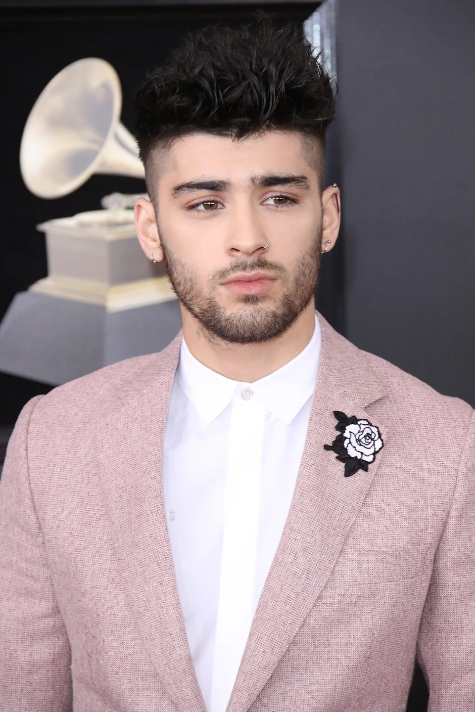 Zayn Malik on red carpet in a stylish suit with floral lapel pin, standing in front of a Grammy Award statue