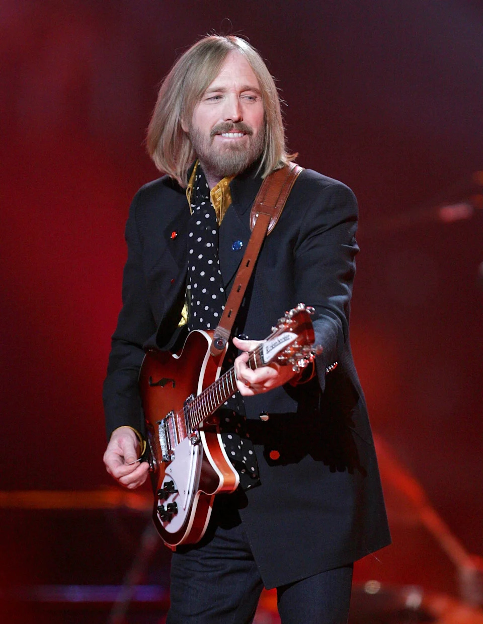A musician performs on stage with a guitar, wearing a patterned shirt and a dark blazer
