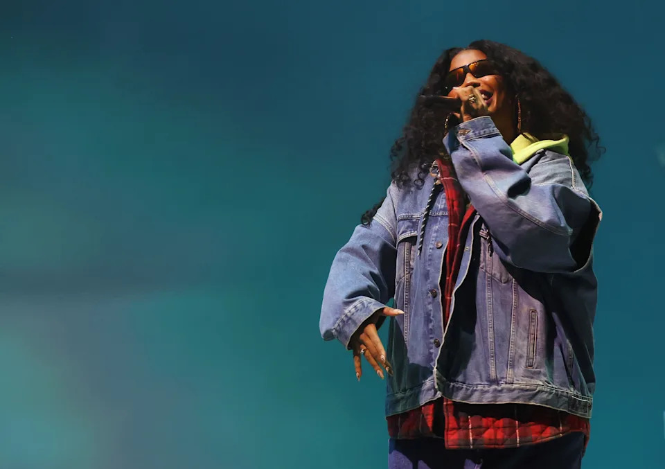 SZA on stage wearing oversized denim jacket, plaid shirt, and sunglasses, performing passionately