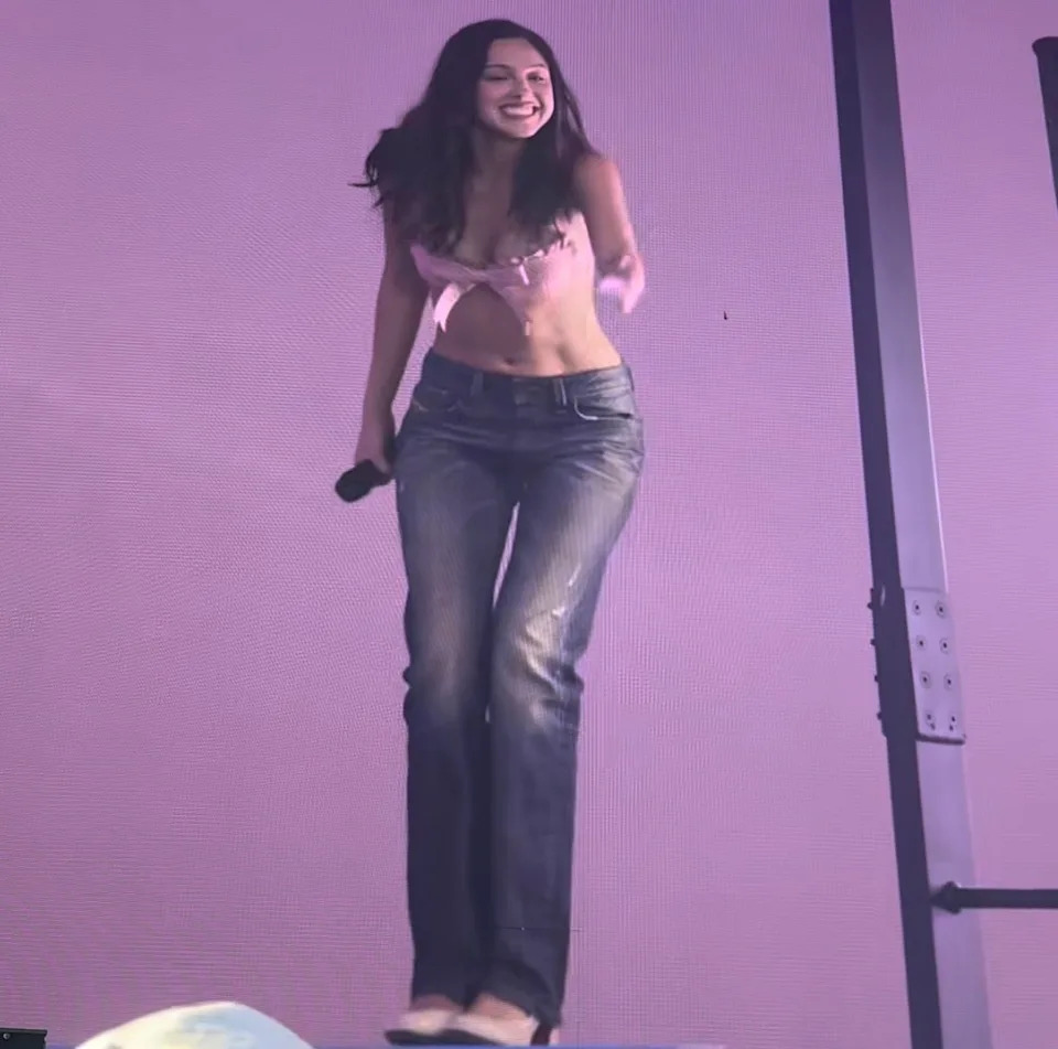 Olivia Rodrigo performs on stage, wearing a stylish top and jeans, holding a microphone and smiling energetically